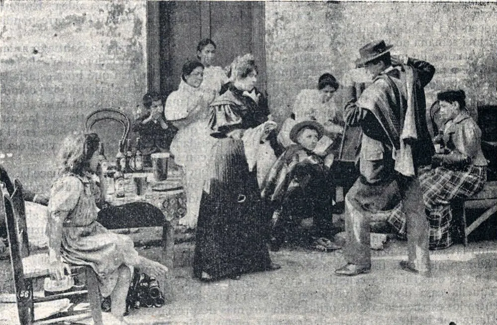 Fotografía de 1900 de una pareja bailando cueca con pañuelos frente a un grupo de personas sentadas.