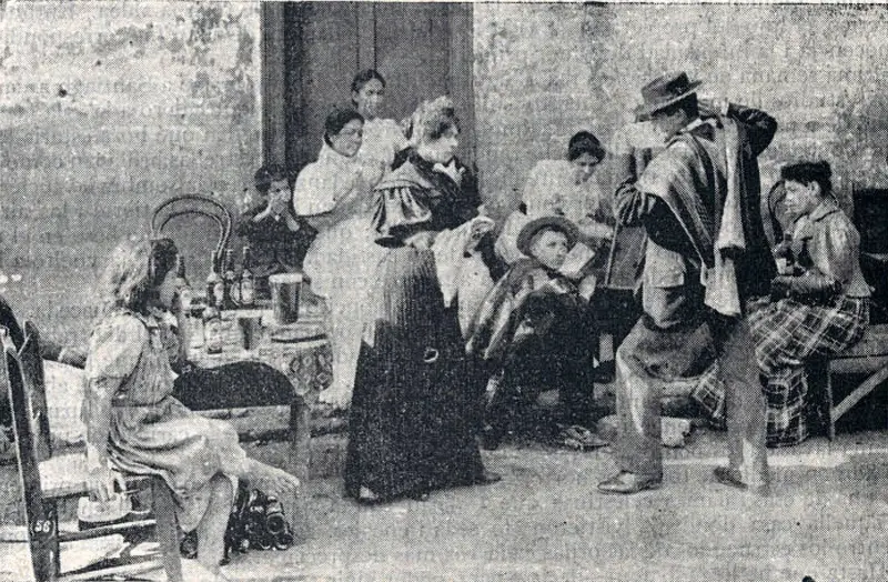 Fotografía de 1900 de una pareja bailando cueca con pañuelos frente a un grupo de personas sentadas.