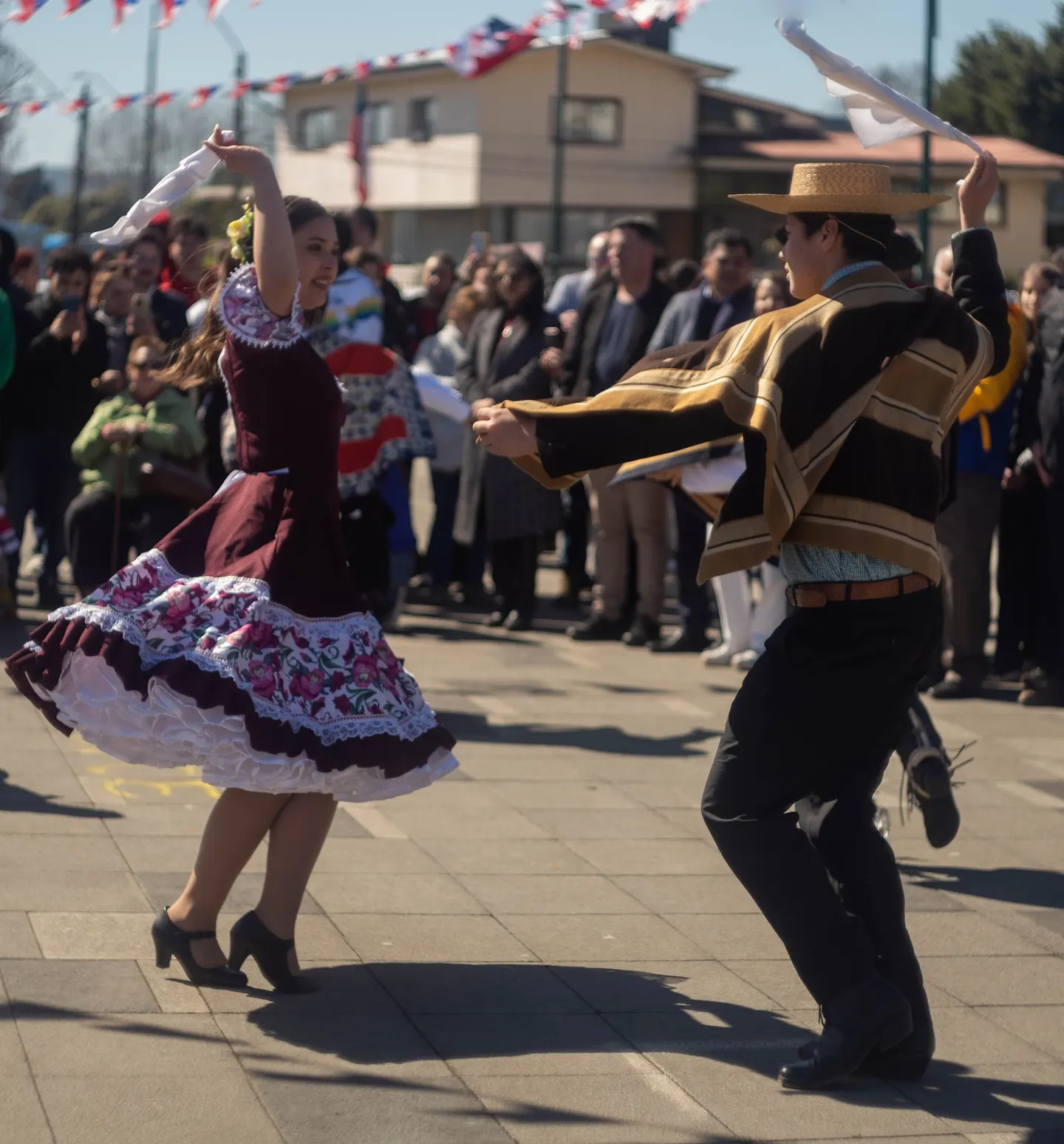 Pareja bailando cueca en una plaza: ambos con el pañuelo en alto, ella con vestido de china floreado y él con manta de huaso.