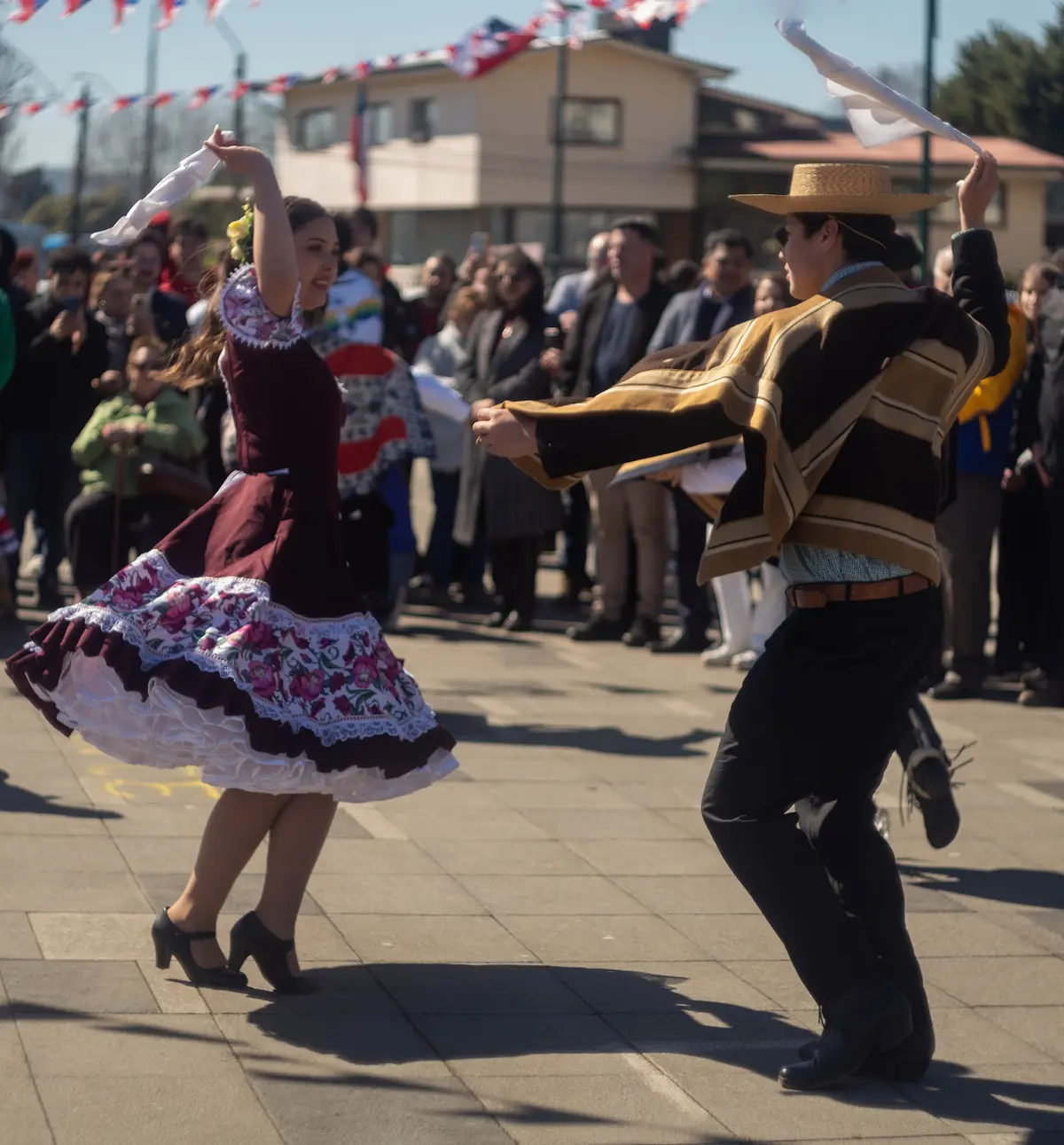 Pareja bailando cueca con los dos pañuelos en alto mientras avanzan por el círculo, ella con vestido burdeos y él con manta.