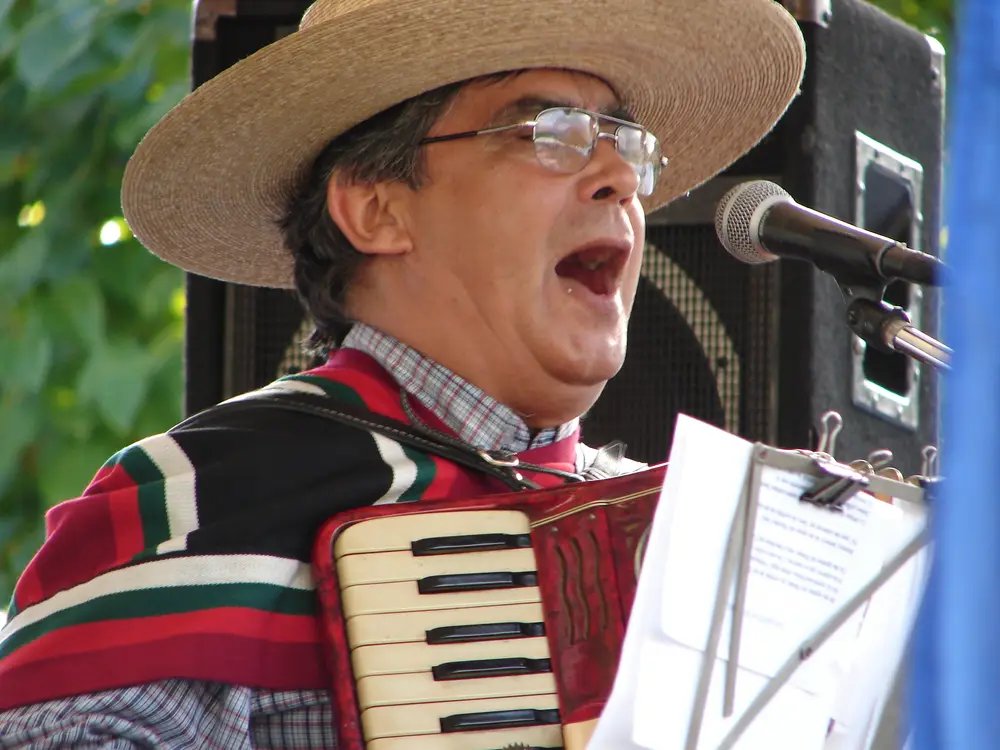 Músico con chupalla y manta cantando y tocando acordeón durante una celebración de Fiestas Patrias.
