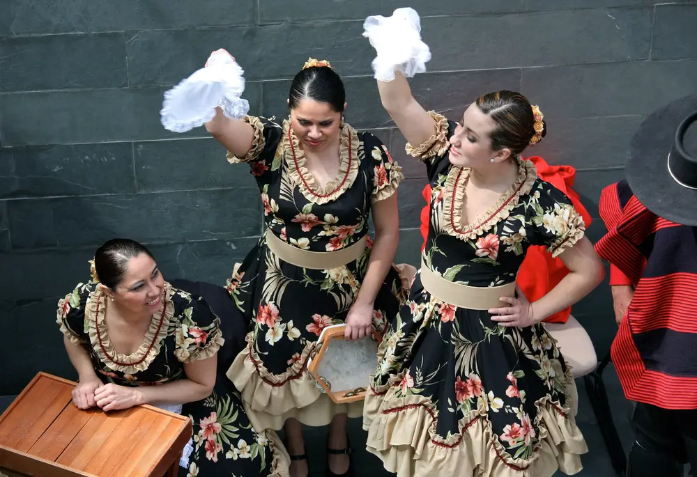 Tres bailarinas de cueca con vestidos floreados levantan el pañuelo por sobre la cabeza, con la muñeca girada.