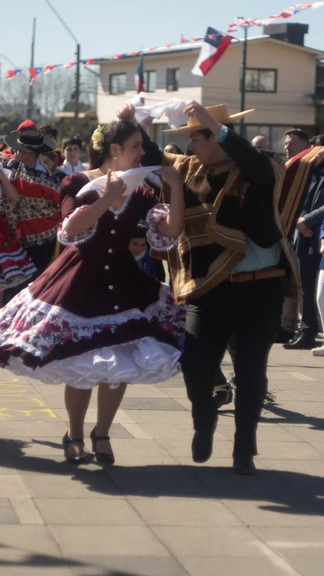 Pareja de cueca huasa muy cerca uno del otro, pañuelos al hombro: ella con vestido burdeos y él con chupalla y manta.