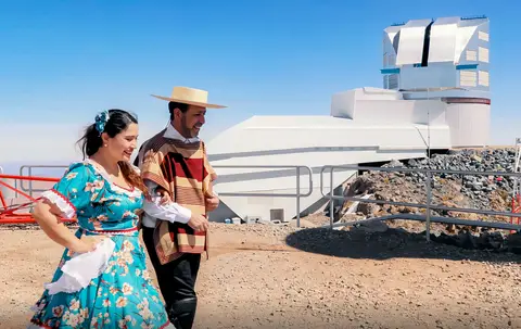 Pareja de cueca caminando del brazo: ella con vestido floreado turquesa y él con chupalla y manta, ambos con el pañuelo en la mano libre.