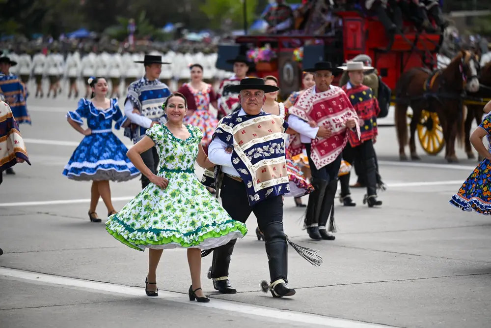Pareja de cueca zapateando de frente: ella con vestido floreado verde y él con manta y espuelas, el taco levantado.